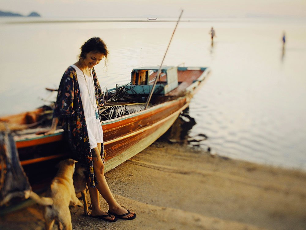 A person standing next to a wooden long-tail boat on a beach at twilight